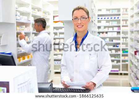 Medium shot of a female pharmacist with a medicine pot and prescription standing at the counter and smiling at the camera.
