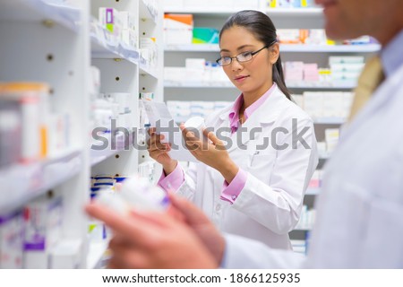 Medium shot of a female pharmacist reading the label on a medicine pot at a pharmacy with a colleague in the foreground.