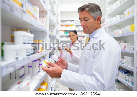 Male pharmacist reading the label on a medicine box at a pharmacy with a colleague in the background.