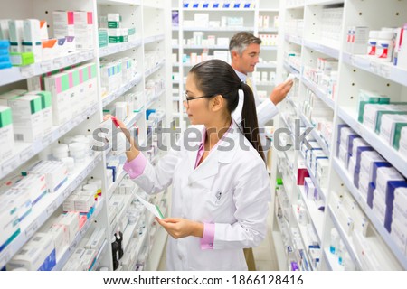 Pharmacist with a prescription looking at a medicine box on a pharmacy shelf.