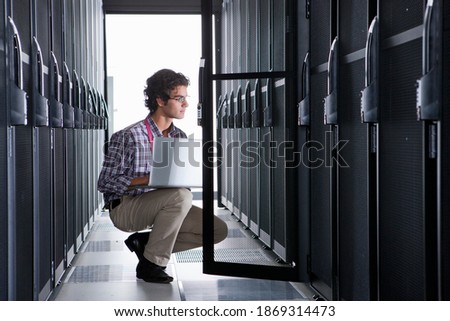 Young technician using a laptop while kneeling in the aisle of a server room.
