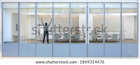 Wide shot through an office window with a businessman standing in the meeting room with his arms raised in the air.