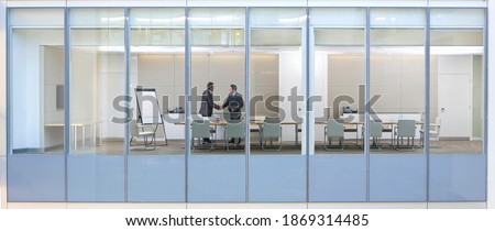 Wide shot through an office window with business men shaking hands in the meeting room.
