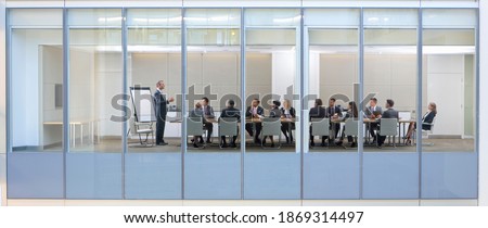 Wide shot through an office window with a businessman in a meeting with his employees.