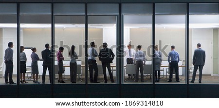 Wide shot through an office window with a group of people standing in front of their manager.