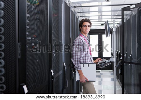 Medium shot of a young technician smiling at the camera while working on a computer connected to a cabinet in the server room.