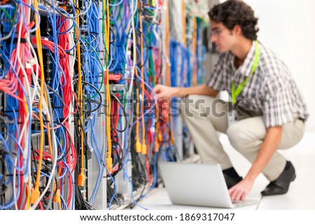 Young technician kneeling next to a cabinet and working on a laptop in a server room with an array of wires in the foreground.