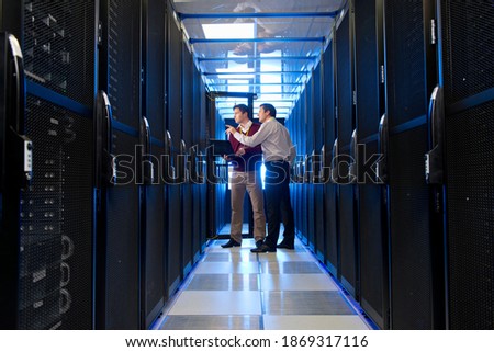 Wide shot of a manager and technician with a laptop having a discussion in the aisle of a server room.
