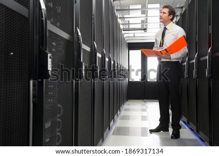 Full shot of a manager standing in the aisle of a data center with a file.