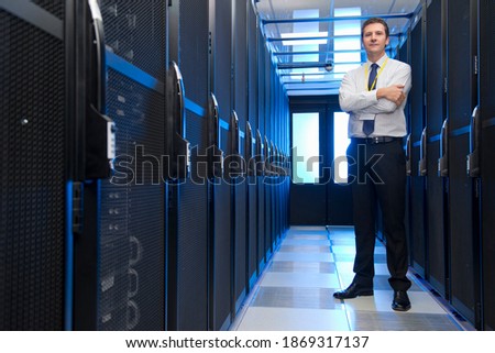 Full shot of a manager with arms crossed standing in the aisle of a data center aligned with storage cabinets.