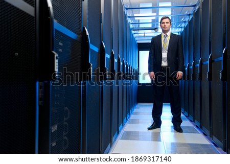 Full shot of a manager in a suit standing in the aisle of a data center aligned with storage cabinets.