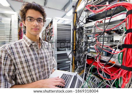 Technician looking at the camera while working on a laptop in a server room at a data center.