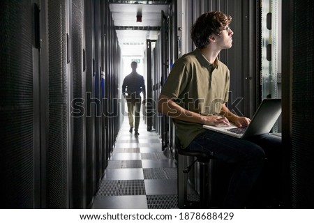 Wide shot of a technician checking the data center with his laptop and his colleague is walking on the floor