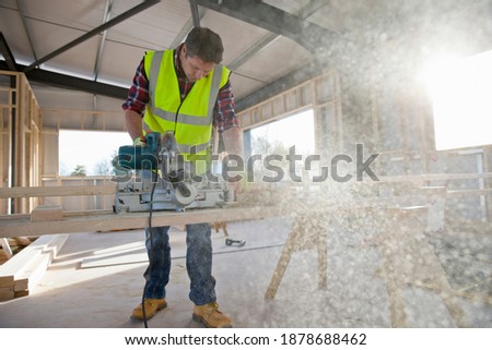 Carpenter is cutting wooden planks at a construction site in a building using a circular blade saw wearing a high visibility safety vest