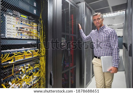 Portrait Of a Technician With Laptop standing In the secured data center leaning on the server