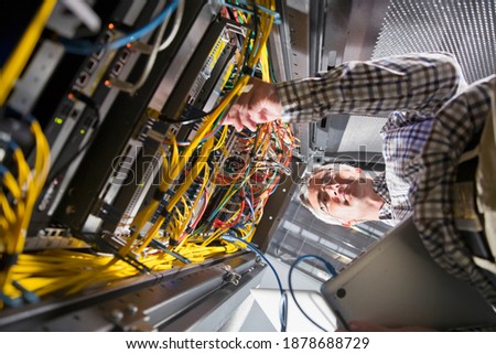 Wide shot of a technician checking the wires in the data center holding a laptop