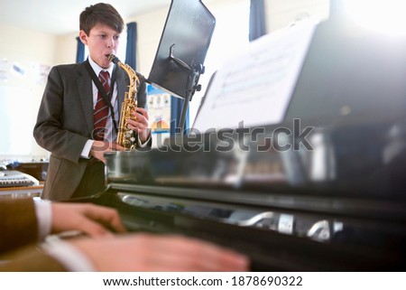High school student reading sheet music and playing a saxophone with a music teacher playing piano in the foreground.