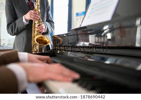 High school student playing a saxophone with a music teacher playing piano in the foreground.