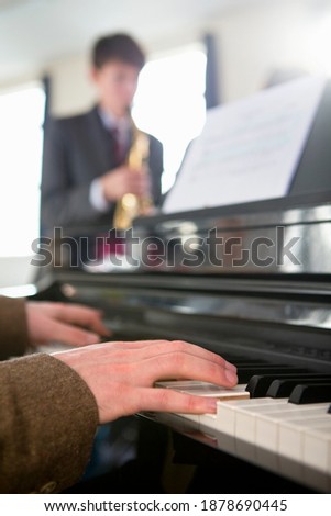 Vertical close-up of a music teacher playing piano with a high school student playing a saxophone in the background.
