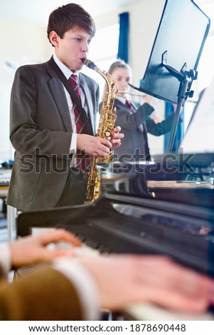 Vertical shot of a high school student reading sheet music and playing a saxophone with a music teacher playing piano in the foreground.
