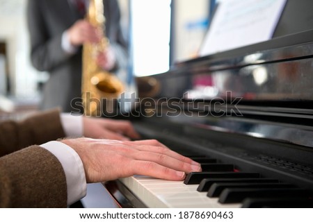 Close-up shot of a music teacher playing piano with a high school student playing a saxophone in the background.