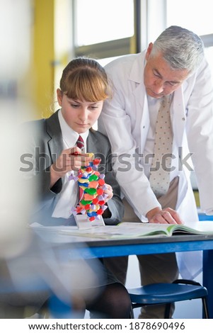 Vertical shot of a science teacher watching a high school student assemble a helix DNA model.