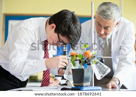 High school student using a microscope during a biology class with a teacher standing next to him.