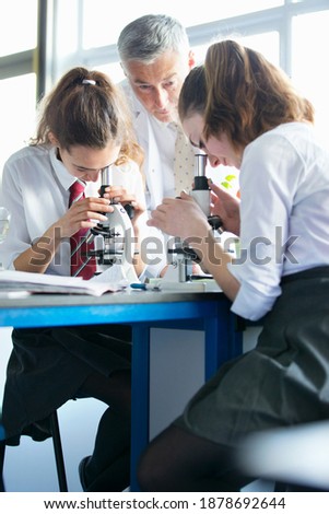 Vertical shot of high school students looking through microscopes in a biology class with a teacher watching them.