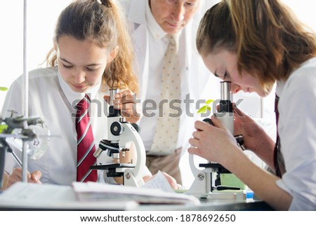 High school students looking through microscopes and making notes in a biology class with a teacher watching them.