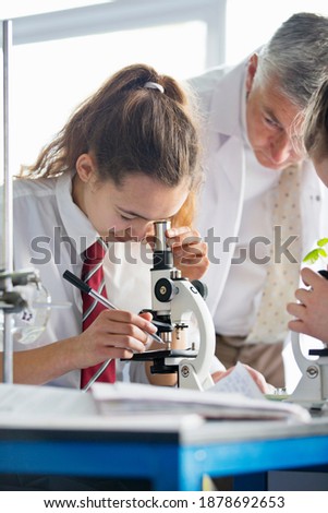Vertical shot of a high school girl looking through a microscope in a biology class with a teacher watching her.