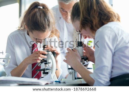 High school students looking through microscopes in a biology class with a teacher watching them.
