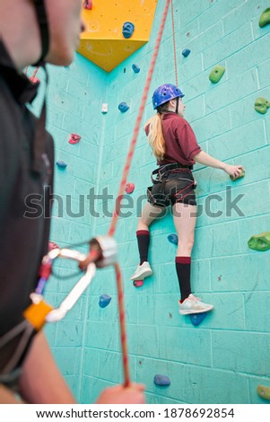 Gym teacher holding the harness rope and watching a high school student rock climbing a wall.