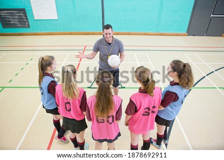 High-angle shot of gym teacher with a netball talking to high school students in a gym.