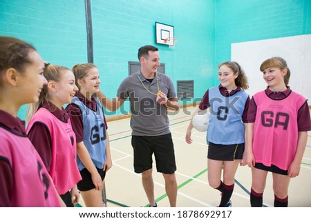 Gym teacher with a whistle talking to high school students with a netball in a gym.