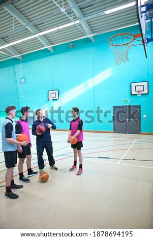 Vertical wide shot of a gym teacher teaching basketball to a group of high school students in a gym.