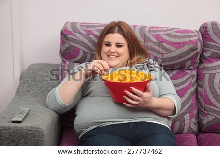 Smiling woman with overweight eating cheese puffs on the couch