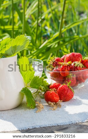 Bowl of strawberries, a cup of milk, a pitcher of strawberry leaves on a napkin on a wooden table on a background of green