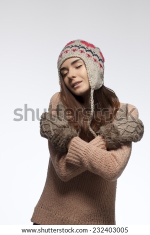 Beautiful young woman in a warm hat and a light brown gloves hugging herself hands on a white background.