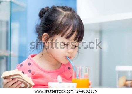 Asian girl sucking juice from a glass in one hand and bread.