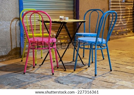 A set of four colored chairs and a small table set on the street.  On the table there is an ashtray and a small glass with a candle inside.