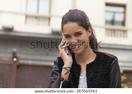 Horizontal shot of a beautiful young woman smiling and talking on the mobile phone.  The picture was taking outdoors with a building in the background in a city.