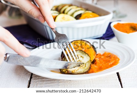 eat warm ratatouille in a round plate with a fork and knife, hand on a white background