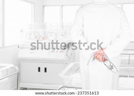 rear view image of doctors with stethoscope looking at newborn baby covered inside incubator in hospital post-delivery room