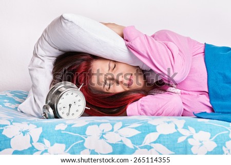 This alarm really annoys me. Young girl in bed with a pillow covering her ears due to alarm ringing