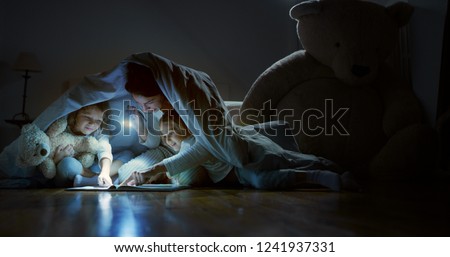 A mother tells stories to her daughters in the dark illuminating with a torch under the blanket. Concept: Love, Family, Dreams
