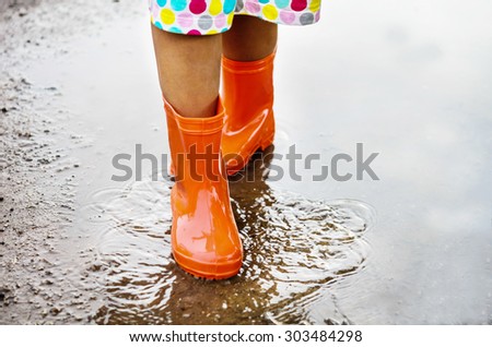 Child wearing orange rain boots walking into a puddle. Close up