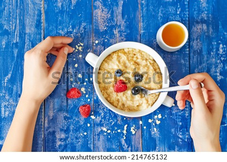Top view showing hands eating porridge with honey and berries on a blue vintage wooden table