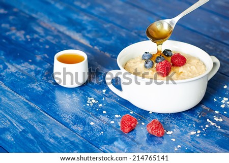 Breakfast with porridge and spoon poring honey, berries, on a blue wooden table
