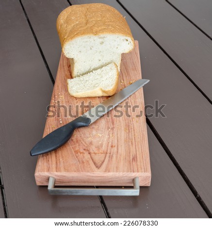 A loaf of white bread on a wooden bread board with a bread knife. A slice of bread has been cut. The background is wooden table slats./Bread Slice/Bread Board