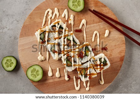 Uramaki Sushi Rolls Topped with White Spicy Cheese Sauce and Brown Soy Sauce Top View. Nori Maki Set on Natural Wooden Background with Thin Slices of Cucumbers and Chopsticks